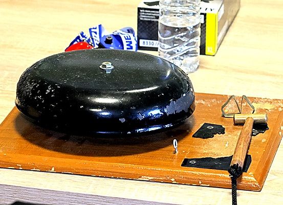 Eine schwarze Glocke steht auf einem Holzbrett mit einem Hammer, neben einer Wasserflasche und einer Schachtel auf einem Holztisch.