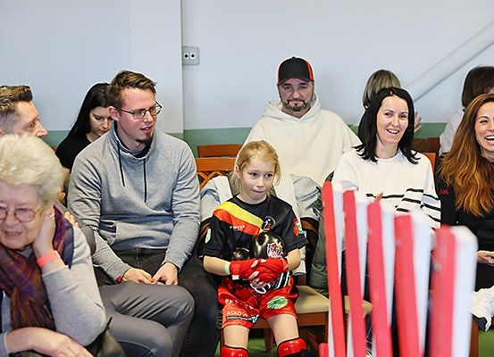 Eine Gruppe von Menschen sitzt in Stühlen in einem Raum, eine junge Mädchen in Sportkleidung schaut nach vorne. Hinter ihr sitzen mehrere Erwachsene, einige lächeln. Im Hintergrund sind eine weiße Wand und rote Stangen zu sehen.
