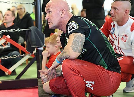 Ein kahlköpfiger Mann mit Tätowierungen kniet in einem Boxring, trägt ein schwarzes und grünes Trikot mit dem WBC-Logo. Er ist von Zuschauern umgeben, darunter ein Mann in einem roten und weißen Trikot.