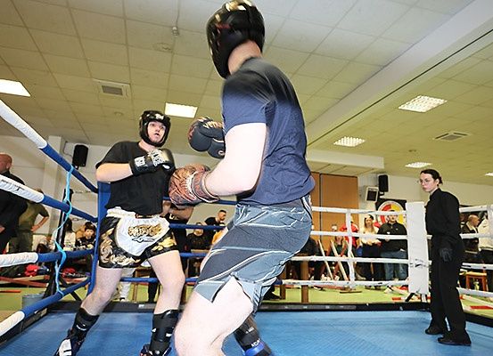 Zwei Männer boxen in einem Ring mit Schutzausrüstung, ein Schiedsrichter steht rechts, und Zuschauer beobachten vom Ringrand aus.