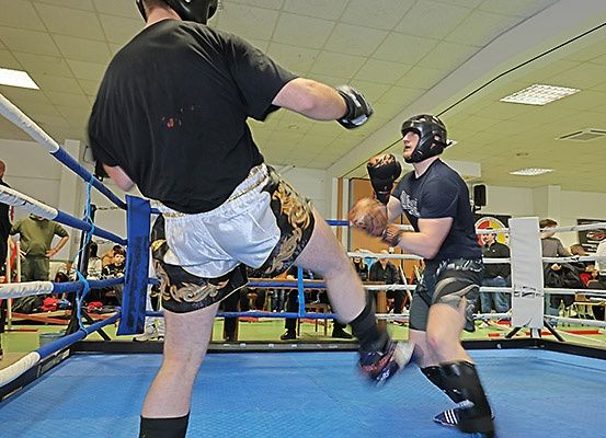 Zwei Männer in einem Boxring, einer trägt schwarze Shorts, beide tragen Helme und Boxhandschuhe, einer schlägt, der andere tritt.