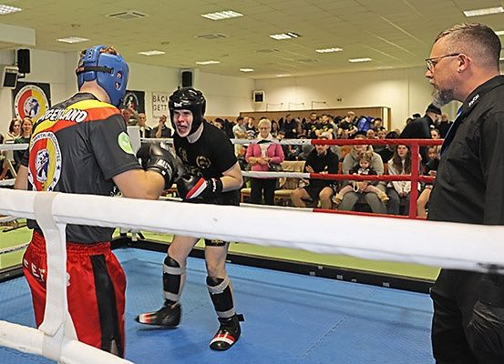 Zwei Boxer sind in einem Ring, einer trägt einen blauen Helm. Ein Schiedsrichter ist anwesend und beobachtet sie. Zuschauer schauen von der Seite zu.