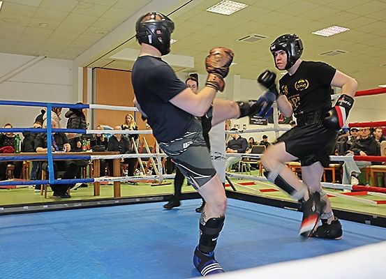 Zwei Männer in einem Boxring mit Helmen und Handschuhen sind in einem Kickboxkampf verwickelt. Sie sind von Zuschauern umgeben.