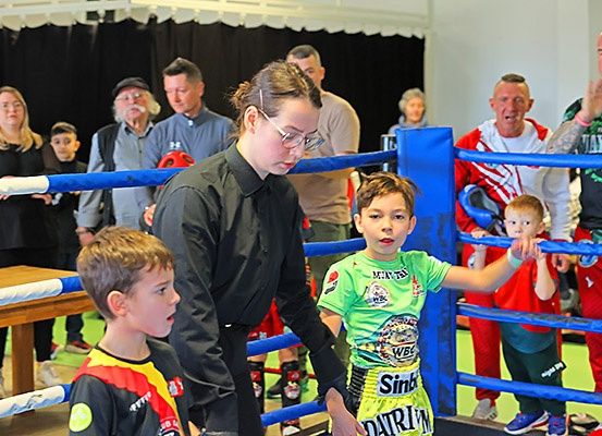 Eine Frau mit Brille führt zwei Kinder, eines in einem grünen Shirt mit dem Wort 'Sin', in einem Boxring, während Zuschauer zusehen.