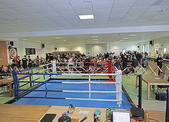 Ein Indoor-Boxring mit einem großen Publikum, das sich um den Ring versammelt hat, um die Veranstaltung zu beobachten. Der Ring hat blaue und weiße Seile, und einige Leute stehen im Inneren.