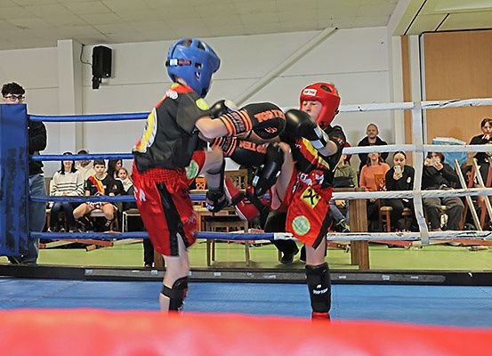 Zwei junge Boxer im Ring, einer in blauer und einer in roter Ausrüstung, sind in einer Kampfhaltung verriegelt. Zuschauer sehen von den Tribünen zu.