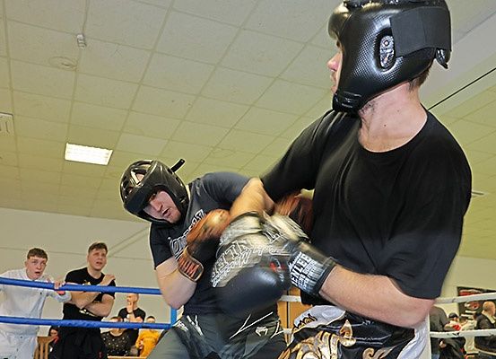 Zwei Männer in einem Boxring üben das Sparring, einer trägt einen schwarzen Helm und Handschuhe, der andere ein schwarzes T-Shirt. Zuschauer beobachten von den Seitenlinien, einige stehen hinter dem Ring.