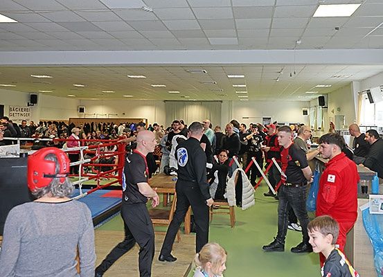 Eine große Gruppe von Menschen in einem Fitnessstudio mit einem Boxring. Mehrere Personen sind in roter und schwarzer Kleidung gekleidet, möglicherweise an einem Wettbewerb teilnehmend.