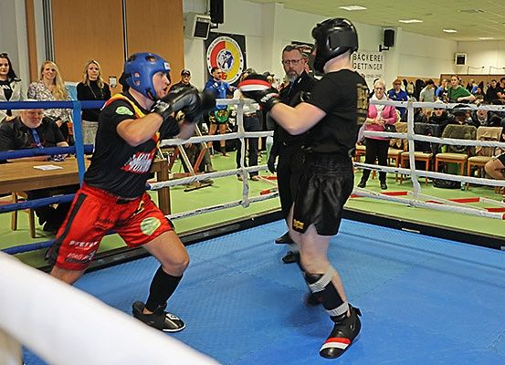 Zwei Boxer im Ring mit Zuschauern. Einer trägt einen blauen Helm, der andere schwarz. Sie tauschen Schläge unter der wachsamen Aufsicht eines Schiedsrichters aus.