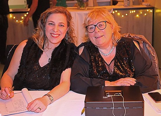 Zwei Frauen sitzen zusammen an einem Tisch, eine schreibt und die andere lächelt. Eine Box mit Kabeln und ein Handy stehen auf dem Tisch. Ein Tisch mit einer weißen Tischdecke und Lichtern steht hinter ihnen.
