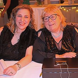 Zwei Frauen sitzen zusammen an einem Tisch, eine schreibt und die andere lächelt. Eine Box mit Kabeln und ein Handy stehen auf dem Tisch. Ein Tisch mit einer weißen Tischdecke und Lichtern steht hinter ihnen.