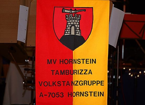 Ein rot-gelber Banner mit einem Wappen zeigt MV Hornstein Tamburizza Volkstanzgruppe A-7053 Hornstein. Der Hintergrund ist dunkel.