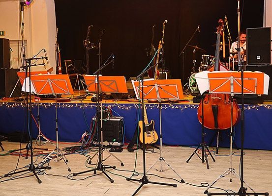 Eine Bühne mit einer Vielzahl von Musikinstrumenten, die für eine Aufführung vorbereitet sind, einschließlich Celli, Gitarren, Trommeln und Mikrofone auf Ständern. Die Bühne ist mit blauem Stoff und Weihnachtslichtern dekoriert.