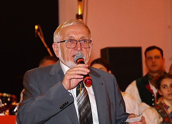 Ein älterer Mann mit Brille und Anzug spricht in ein Mikrofon und hält ein Stück Papier vor einer Menschenmenge.