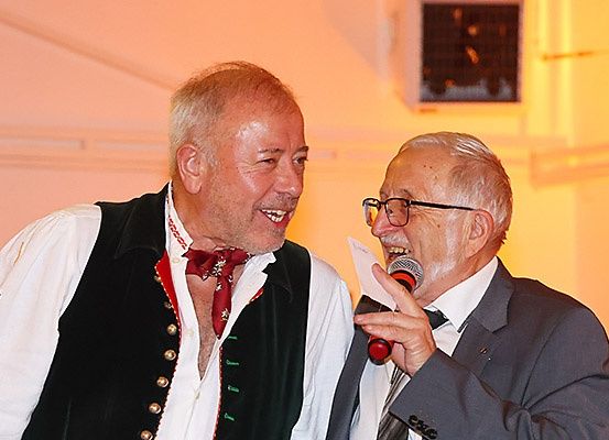 Zwei Männer, einer mit rotem Fliegenbinder und Weste, der andere in Anzug und Brille, stehen lächelnd nebeneinander, einer hält ein Mikrofon.
