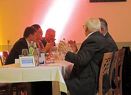 Eine Gruppe von Männern in Anzügen sitzt um einen Tisch in einem Restaurant. Sie unterhalten sich und lachen, und es gibt Flaschen und Gläser auf dem Tisch.