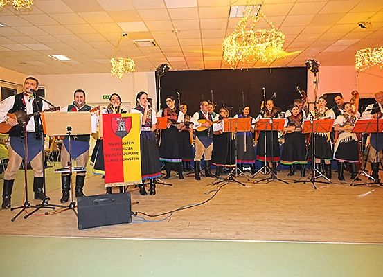 Eine Gruppe Musiker auf der Bühne, einige spielen Gitarren, andere Saiteninstrumente, mit einem Banner im Hintergrund und dekorierten Kronleuchtern.
