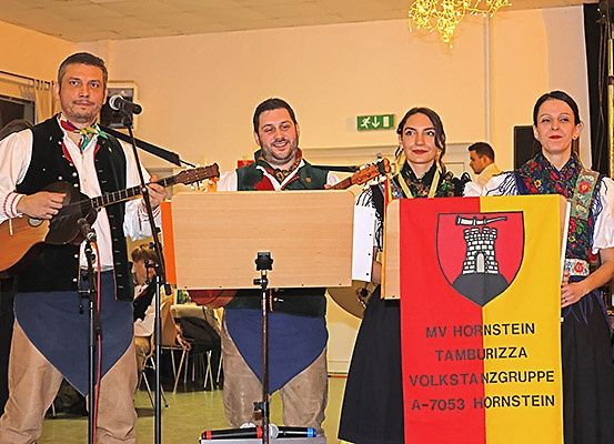 Vier Personen, gekleidet in traditioneller Tracht, spielen Gitarren. Sie stehen vor einem Banner mit dem Wappen einer Burg und dem Namen MV Hornstein Tamburizza Volkstanzgruppe A-7053 Hornstein. Ein Mikrofon ist vor ihnen aufgestellt.