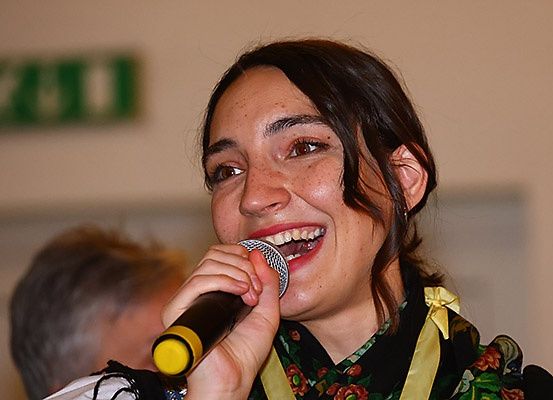 Eine Frau in einem Blumenkleid hält ein Mikrofon, lächelt und singt. Eine Person im Hintergrund ist leicht verschwommen.