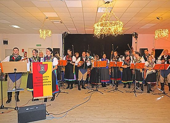 Eine Gruppe von Musikern tritt auf der Bühne in traditioneller Kleidung auf, begleitet von dekorativen Kronleuchtern. Einige spielen Saiteninstrumente, während andere singen. Musikstände sind vor ihnen aufgestellt.