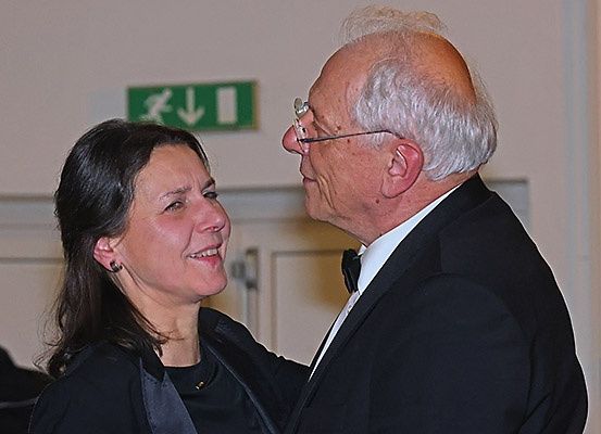 Ein älteres Paar in formeller Kleidung steht eng beieinander, lächelt und unterhält sich möglicherweise bei einer Veranstaltung in Innenräumen.