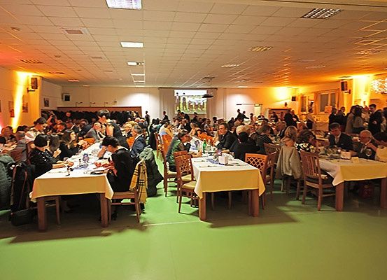 Eine große Gruppe von Menschen sitzt in einem Bankettsaal an Tischen, einige essen und andere unterhalten sich. Der Raum ist mit gelbem Licht beleuchtet und hat eine Projektionsleinwand hinten.