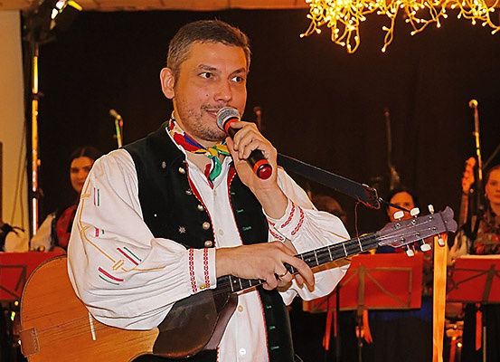 Ein Mann singt in ein Mikrofon, während er Gitarre spielt, umgeben von Musikern mit Notenblättern.