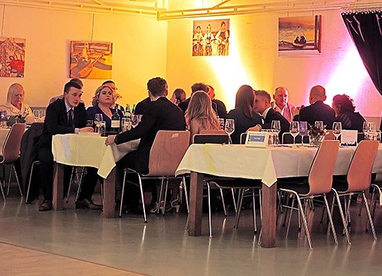 Ein Restaurant mit Tischen und Stühlen, Leute sitzen und unterhalten sich. Jeder Tisch hat Weingläser und Flaschen. Der Raum hat Gemälde an der Wand.