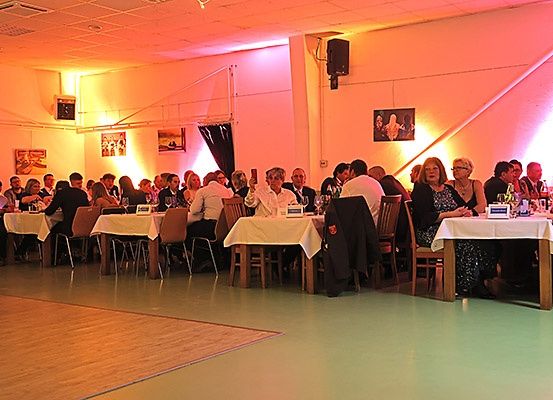 Ein Speisesaal mit mehreren Tischen und Stühlen. Leute sitzen und stehen, einige mit Weingläsern. Es gibt Bilder an der Wand und einen Lautsprecher. Der Raum hat einen Holzboden und pinke Beleuchtung.