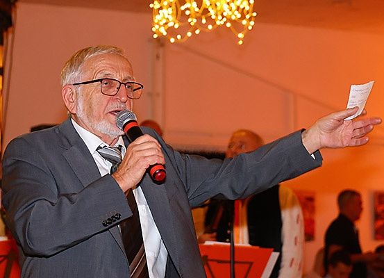 Ein älterer Mann in Anzug und Krawatte spricht in ein Mikrofon und hält ein Stück Papier in der Hand. Hinter ihm stehen mehrere Menschen und schauen zu ihm.