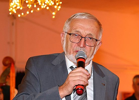 Ein älterer Mann mit Brille spricht in ein Mikrofon bei einer Veranstaltung. Der Hintergrund ist verschwommen, mit Menschen und einer Kronleuchter.
