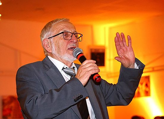 Ein älterer Mann in Anzug und Krawatte hält ein Mikrofon und spricht bei einer Veranstaltung.