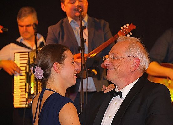 Ein älterer Mann und eine Frau lächeln sich an, während sie eng beieinanderstehen. Hinter ihnen spielt ein Mann Gitarre und ein anderer Akkordeon.