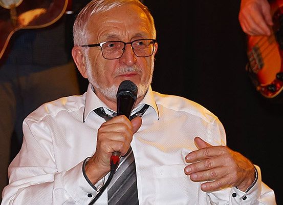 Ein älterer Mann in weißem Hemd und Krawatte hält ein Mikrofon und spricht zu einem Publikum. Hinter ihm spielt eine Person Gitarre.