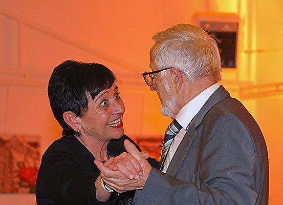 Ein älterer Mann und eine ältere Frau tanzen in einem orange beleuchteten Raum, beide in formeller Kleidung. Die Frau lächelt und hält die Hand des Mannes. Hinter ihnen schmückt ein Kunstwerk die Wand.