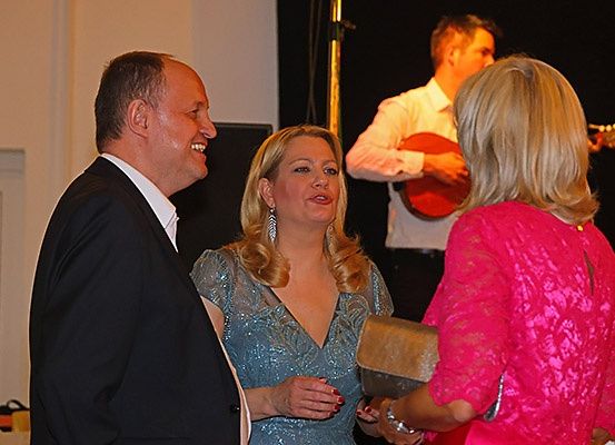 Drei Personen versammeln sich bei einer Veranstaltung, ein Mann in einem schwarzen Anzug, eine Frau in einem blauen Kleid und eine weitere Frau in einem rosa Kleid. Ein Mann spielt im Hintergrund eine Gitarre.
