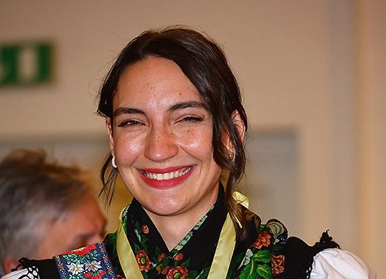Eine Frau mit braunen Haaren, die ein Blumenkleid und einen Schal um den Hals trägt, lächelt. Hinter ihr ist ein verschwommenes Bild einer Person mit grauem Haar. Der Hintergrund ist schlicht mit einem grünen Schild an der Wand.