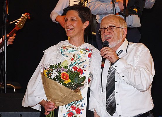 Eine Frau mit Blumen und ein Mann mit einem Mikrofon stehen zusammen auf der Bühne, ein Gitarrist im Hintergrund.