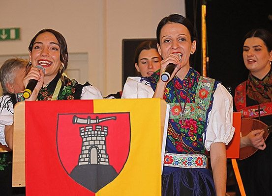 Eine Gruppe von Frauen in traditioneller Kleidung tritt auf, eine hält ein Mikrofon und eine Fahne mit einem Burgwappen. Eine andere Frau spielt hinter ihnen ein Musikinstrument.