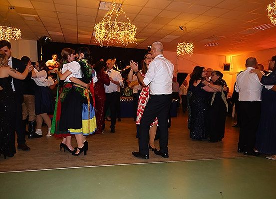 Eine Gruppe von Menschen tanzt auf einer Bühne mit Kronleuchtern darüber. Einige tragen traditionelle Kostüme. Andere tragen formelle Kleidung. Ein Mann spielt Gitarre.