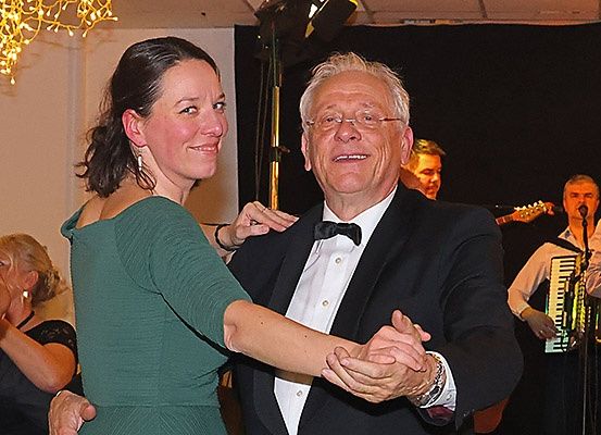 Ein älterer Mann und eine Frau tanzen bei einer gesellschaftlichen Veranstaltung zusammen. Die Frau lächelt und trägt ein grünes Kleid. Hinter ihnen tanzen andere Leute. Im Hintergrund spielt eine Band mit Instrumenten wie Gitarre und Akkordeon.
