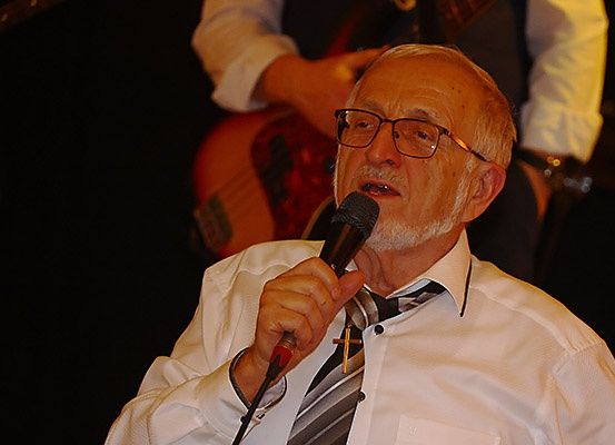 Ein älterer Mann mit Brille hält ein Mikrofon und singt vor einem unscharfen Hintergrund eines Gitarristen.