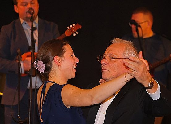 Ein älterer Mann und eine junge Frau tanzen auf der Bühne, während eine Band im Hintergrund spielt.