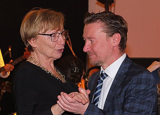 Eine ältere Frau mit Brille und einer Halskette tanzt mit einem Mann in einem Anzug bei einer gesellschaftlichen Veranstaltung.