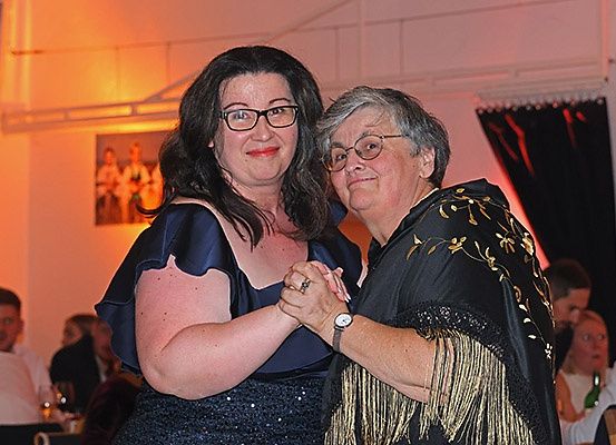 Zwei Frauen, eine in einem blauen Kleid und die andere in einem schwarzen Kleid mit Goldstickerei, tanzen zusammen in einem Raum mit gedämpfter Beleuchtung und mehreren Personen, die an Tischen sitzen.