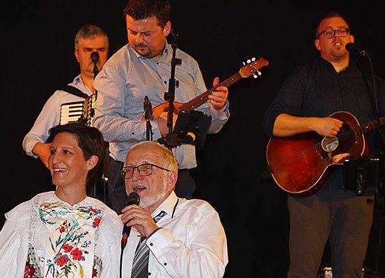 Eine Band tritt mit einer Frau und einem älteren Mann auf der Bühne auf. Der Mann spielt Gitarre und singt in ein Mikrofon, während ein anderer Akkordeon spielt. Ein Dritter spielt Gitarre.