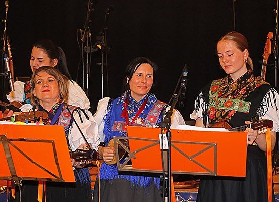 Vier Frauen in traditioneller Kleidung spielen Gitarren, stehen vor orangefarbenen Notenständern auf der Bühne, mit dunklem Hintergrund.
