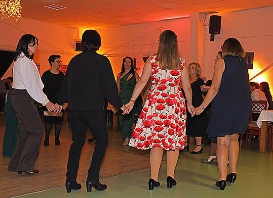 Mehrere Frauen tanzen in einem Raum mit gedämpfter Beleuchtung und halten sich an den Händen. Eine Frau in einem Blumenkleid steht in der Mitte. Eine Kronleuchter und ein Lautsprecher sind im Hintergrund.