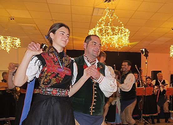 Ein Mann und eine Frau tanzen in traditioneller Kleidung inmitten einer festlichen Umgebung, während Musiker verschiedene Instrumente spielen.