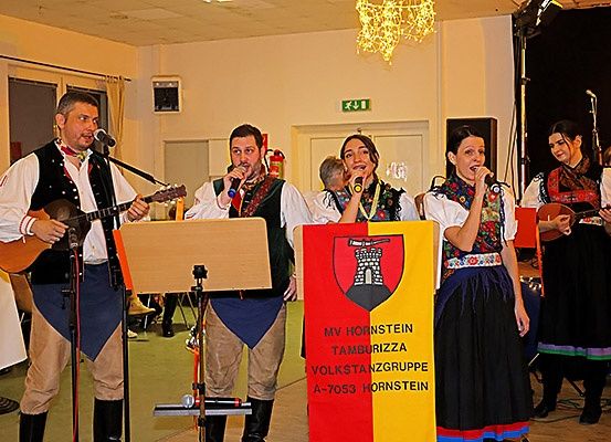 Eine Gruppe von Musikern tritt auf der Bühne auf, wobei zwei Frauen und zwei Männer in Mikrofone singen. Der Mann auf der linken Seite spielt Gitarre, während ein Banner ein Logo und Text zeigt.